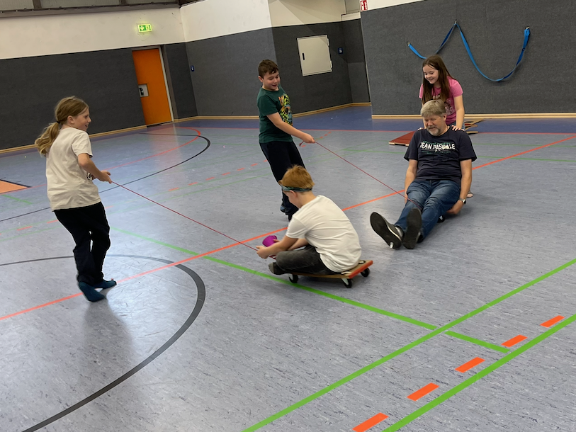 Kinder ziehen einen Erwachsenen auf einem Rollbrett in einer Turnhalle