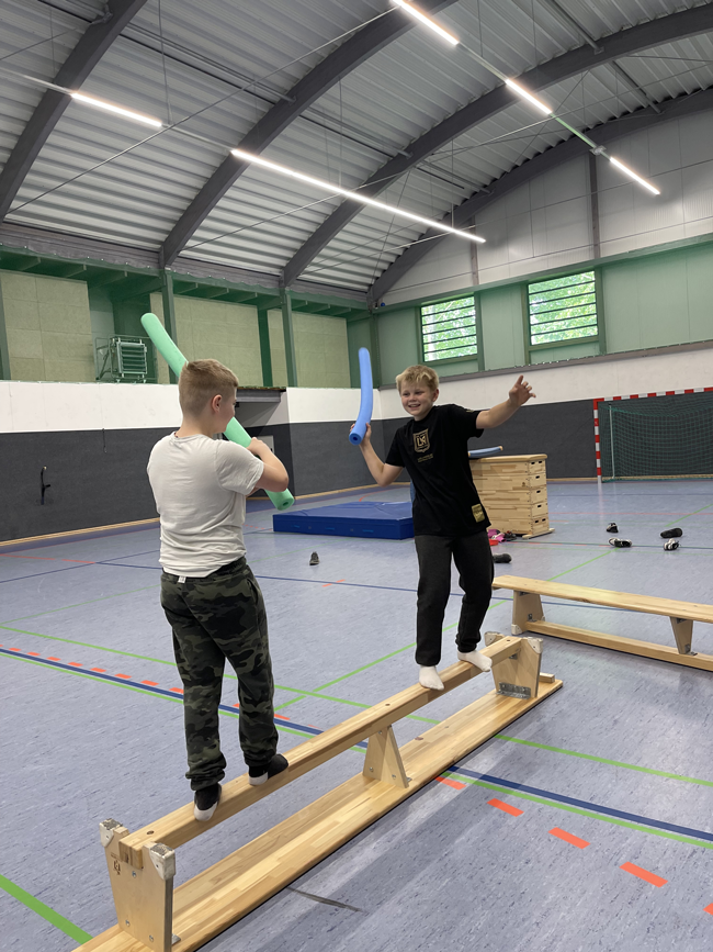 Zwei Jungen duellieren sich mit Poolnudeln auf einer umgedrehten Bank 