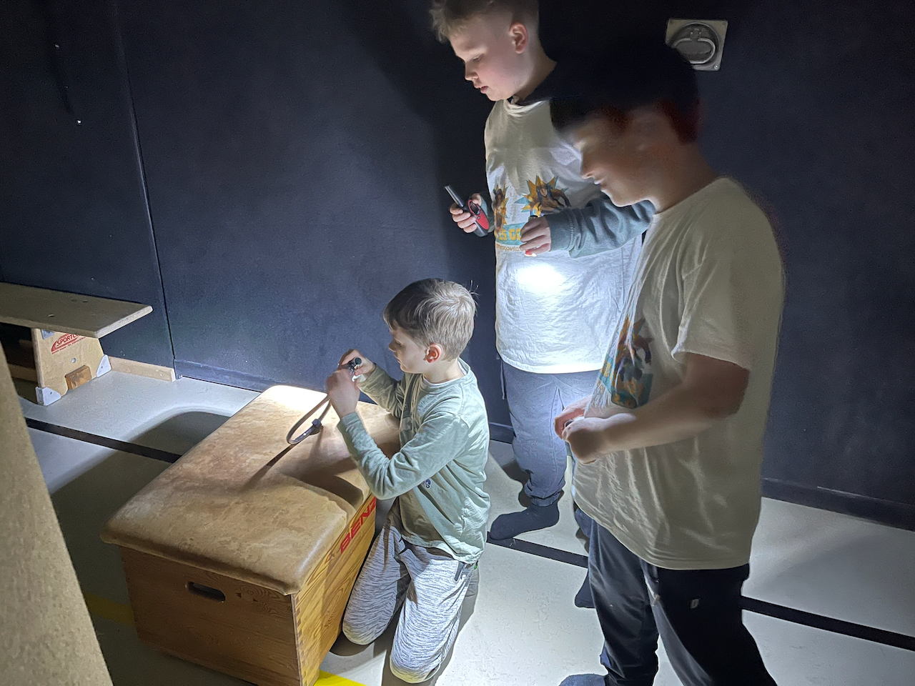 Mehrere Jungs öffnen ein Zahlenschloss in einer dunklen Turnhalle, sie geben sich Licht mit Taschenlampen.