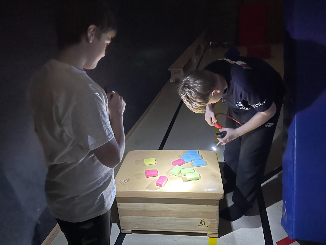 In einer Turmhalle: Im Dunklen wird ein Zahlenschloss von einem Jungen geöffnet, während ein anderer ihm Licht mit einer taschenlampe gibt.