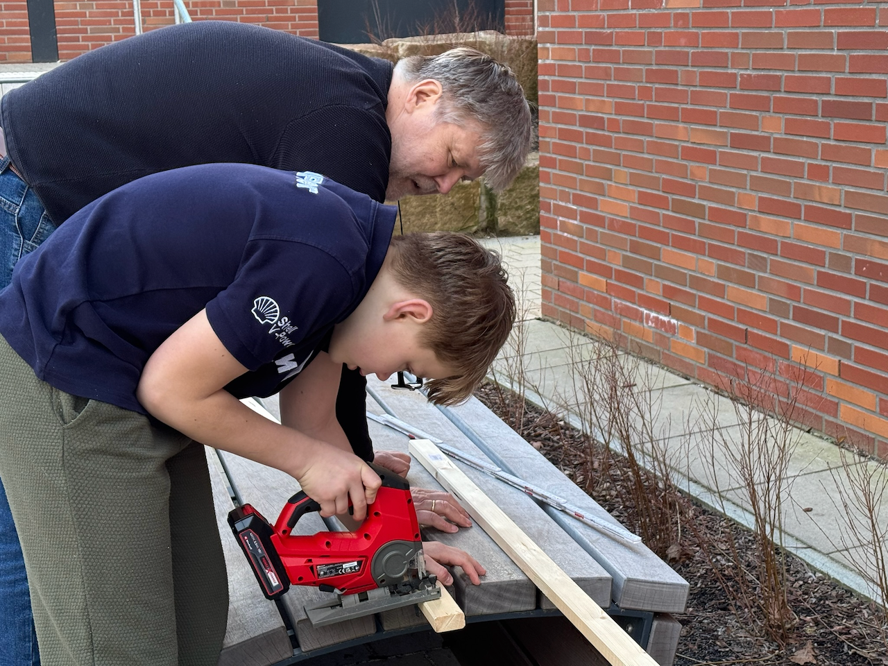 Junge sägt mit Stichsäge Leiste ab, Mann dahinter hält das Holz fest.